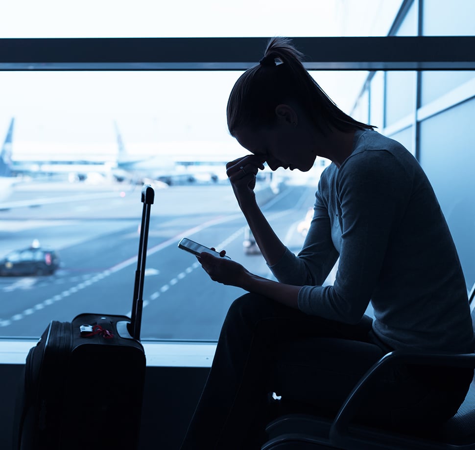 Eine Frau die mit einem Koffer bei einem Flughafen sitzt. Im Hintergrund sind durch ein Fenster Flugzeuge zu sehen.