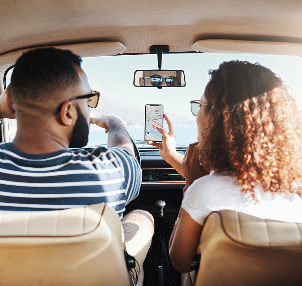 Zwei Personen sitzen in einem Auto. Die Personen sind von hinten zu sehen. Die rechte Person zeigt der linken Person die fährt eine Navigations-App auf einem Smartphone. 
